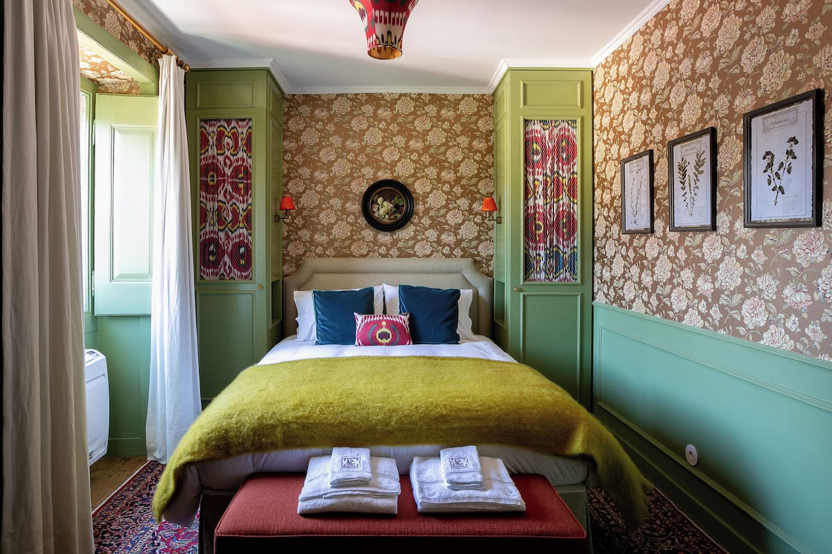 A neatly arranged bedroom with a bed, pillows, a bedside table with towels, and a wall-mounted clock.