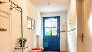 A narrow hallway with a blue door, a bench with red cushions, a radiator, and a wall-mounted shelf with a plant. - Papillon Wohnung 15-7 (Ostseebad Boltenhagen)