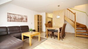 A modern living room with a sofa, a wooden coffee table, and a dining area with chairs and a table. There is a wooden staircase and a bookshelf in the background. - Papillon Wohnung 15-7 (Ostseebad Boltenhagen)