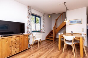Ein Zimmer mit einem Holzschrank, einem Fernseher, einem Esstisch mit Stühlen, einer Treppe und einem Fenster mit Vorhängen.