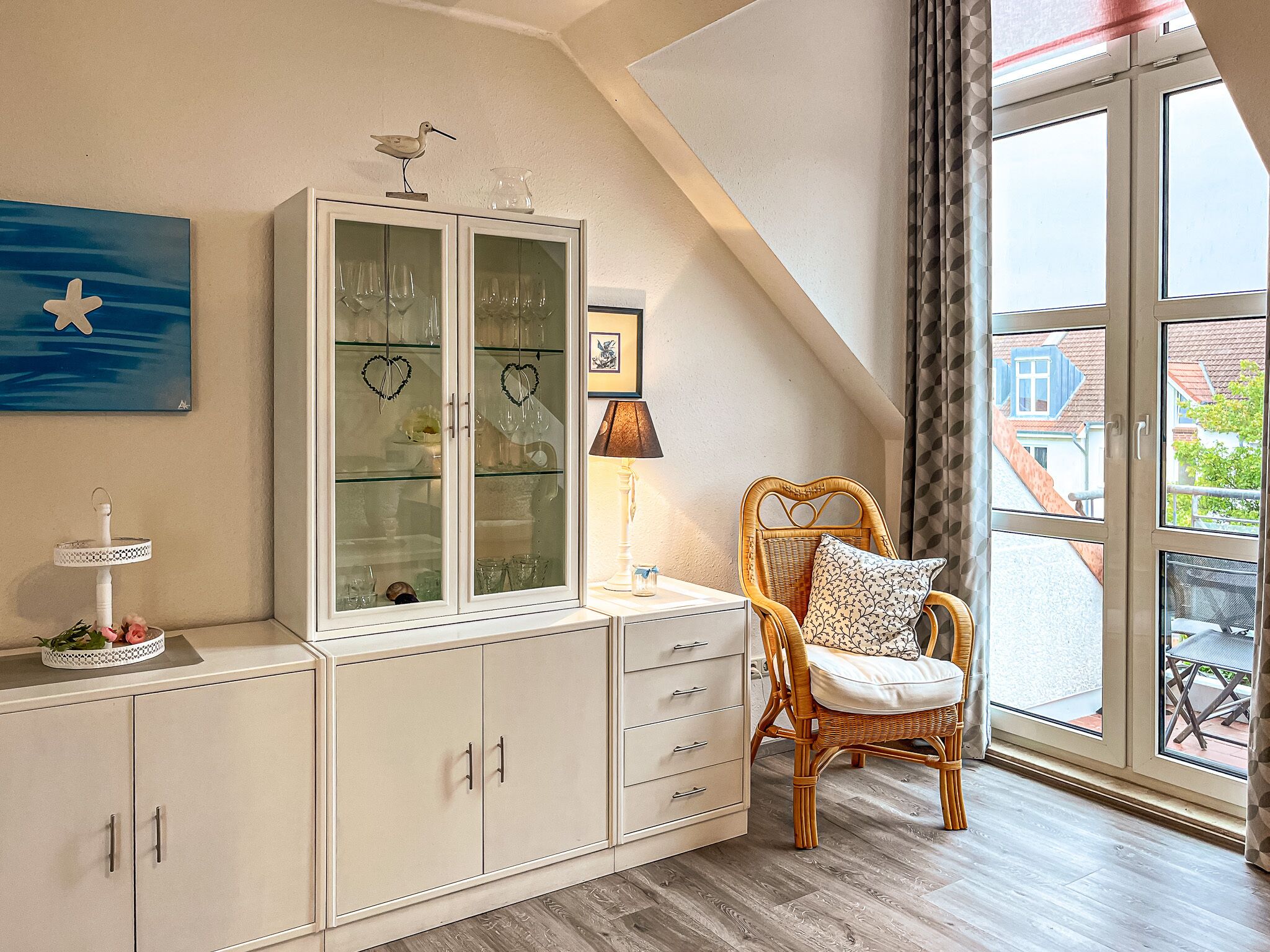 Une pièce avec un meuble blanc aux portes vitrées, une chaise en osier avec un coussin, une lampe et une vue par la fenêtre.
