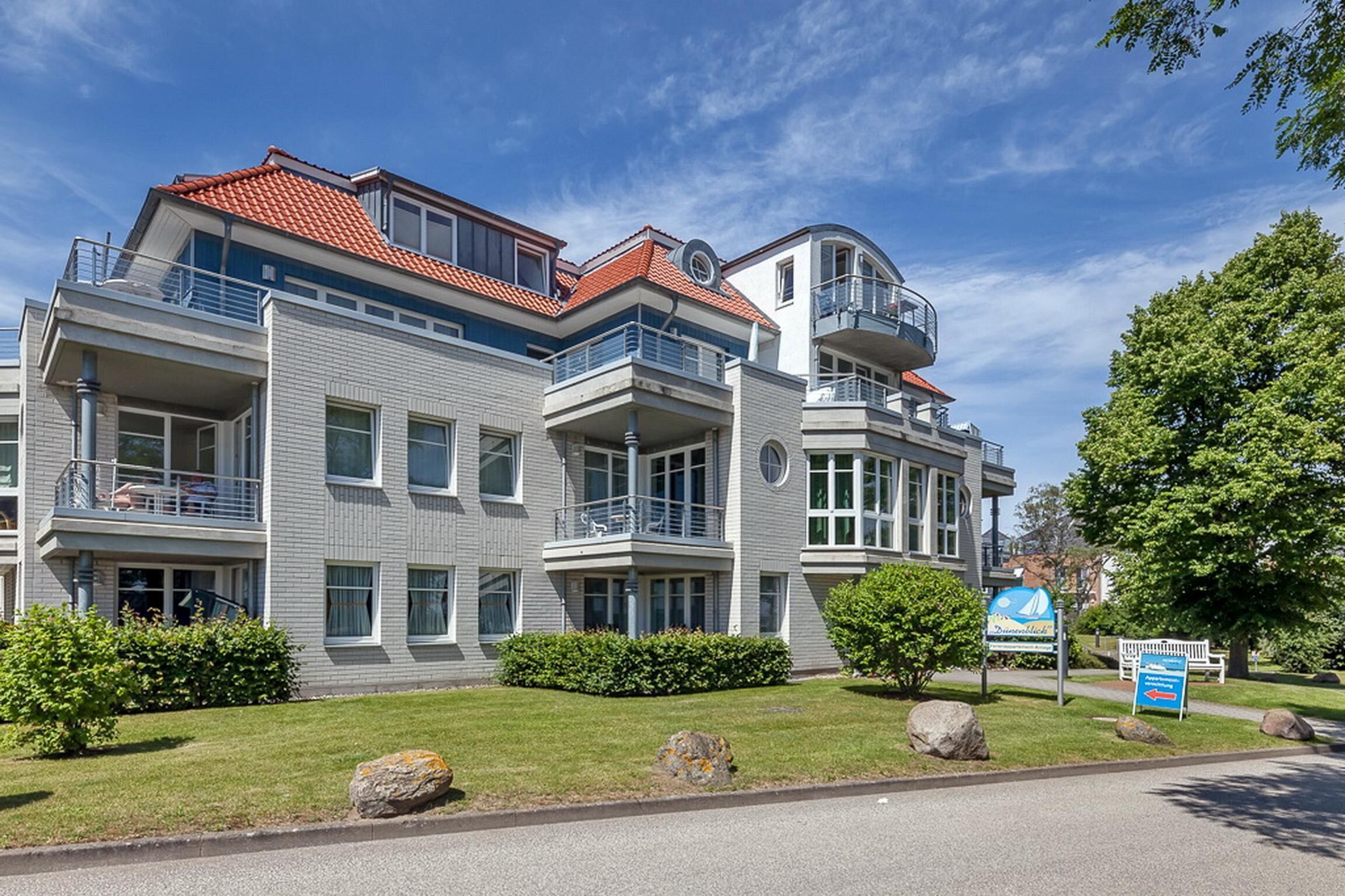 A multi-story residential building with balconies, a red-tiled roof, and a well-maintained lawn with rocks and bushes.