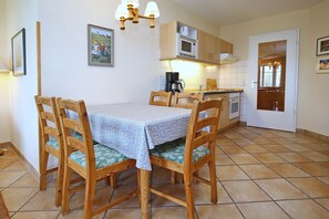 Un coin repas avec une table dressée pour quatre personnes, des chaises en bois dotées d’assises rembourrées vertes, un luminaire en bois au plafond, et un espace cuisine avec un four à micro-ondes et une cuisinière en arrière-plan.