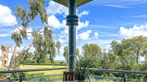 Een houten tafel met een potplant op een balkon met een hekwerk, met uitzicht op een gazon en bomen.