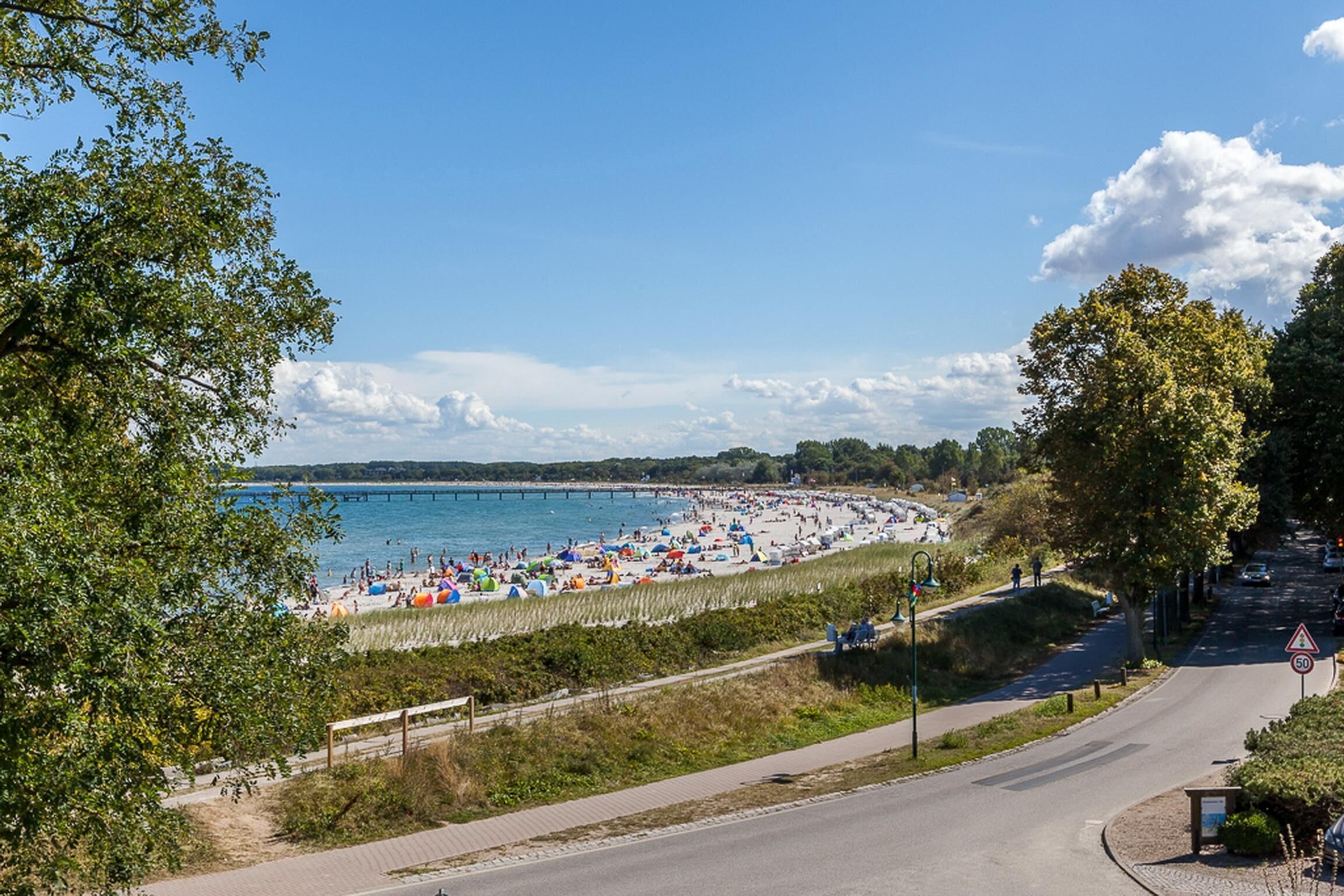 Uma praia com um calçadão pavimentado, uma estrada que curva para a direita e um píer que se estende sobre a água.