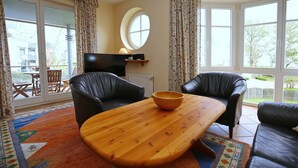 Een woonkamer met een houten salontafel, twee zwarte leren banken, een televisie op een meubel en een groot raam met gordijnen.