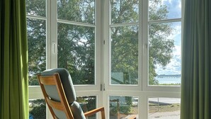 Puinen tuoli, jossa on pehmuste, on sijoitettu suuren, useista ruuduista koostuvan ikkunan eteen. Ikkunasta avautuu näkymä puihin ja vesistöön.