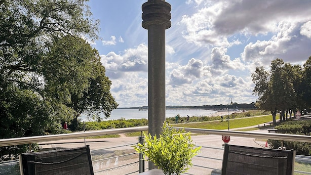 Pöytä, jonka päällä on ruukussa oleva kasvi, ja pöydältä avautuu kaunis näkymä vesistöön ja sen ylle johtavalle sillalle.