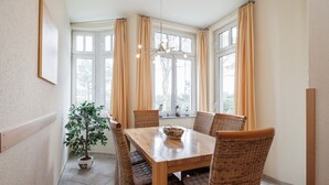 Un comedor con una mesa de madera, sillas de mimbre, una planta en maceta y una ventana con cortinas.