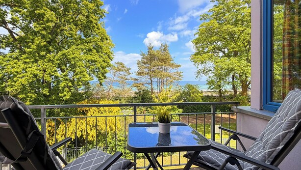 A balcony with two chairs, a small table with a plant, and a view of trees and a body of water. - Villa Triton Wohnung 03 (Ostseebad Boltenhagen)
