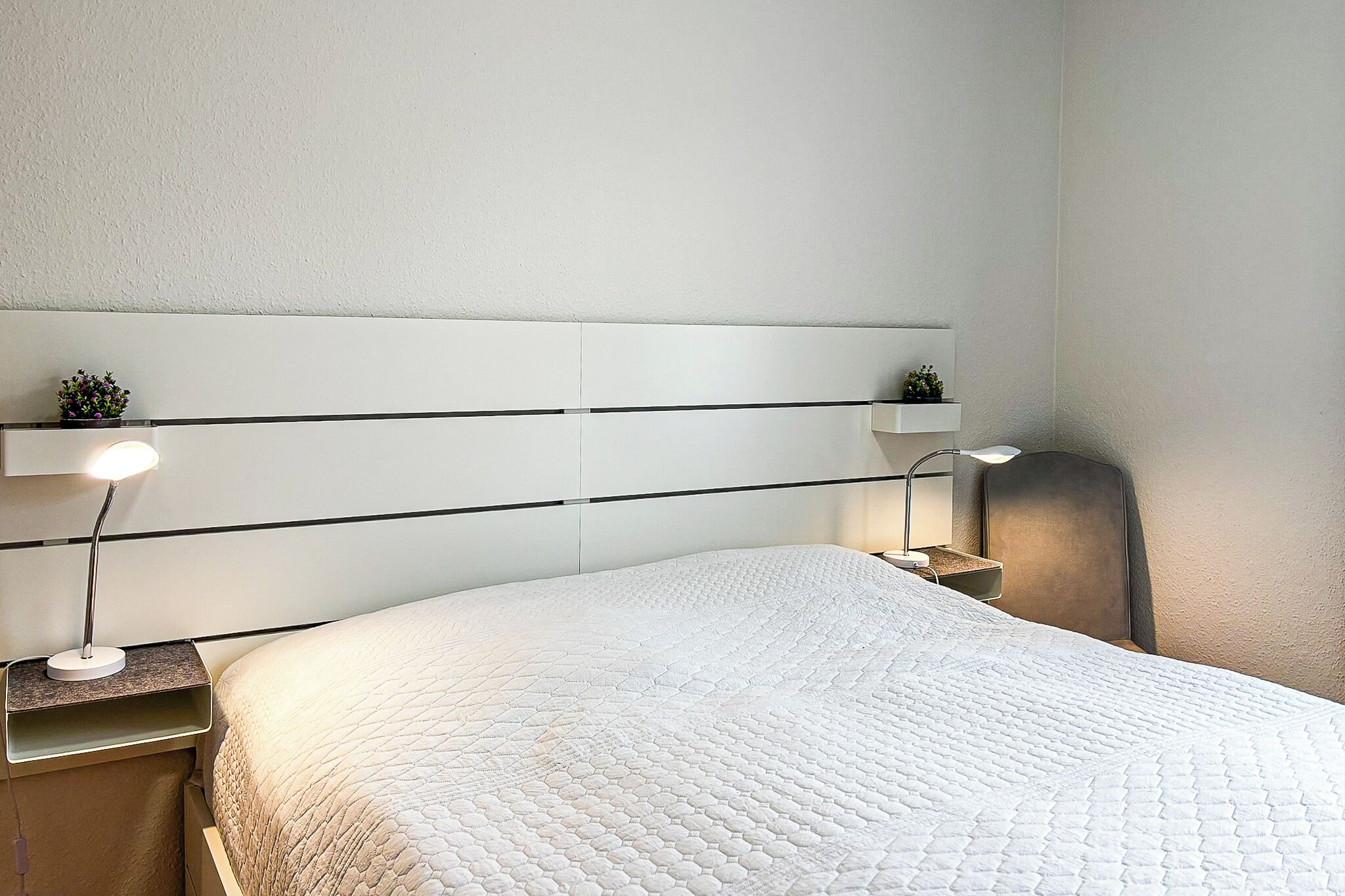 A neatly made bed with a quilted white duvet, a bedside table with a lamp, and a small plant on the nightstand.