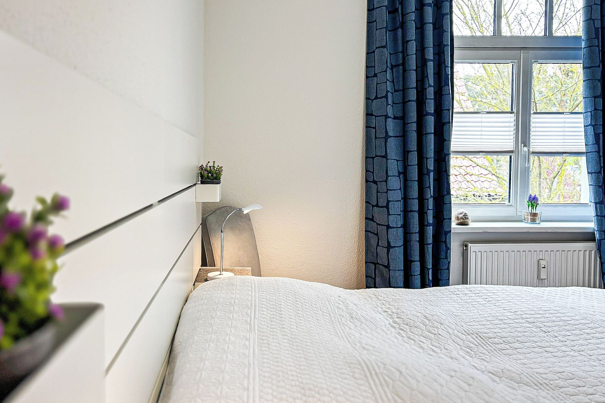 A bedroom with a bed, a bedside table with a lamp, a window with curtains, and a radiator.