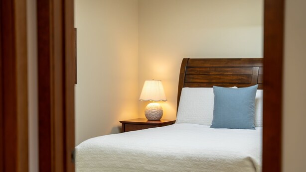 Ein ordentlich bezogenes Bett mit weißer Decke, blauem Kopfkissen, einem Holz-Kopfteil und einer Nachttischlampe mit einer strukturierten Lampenschirm.