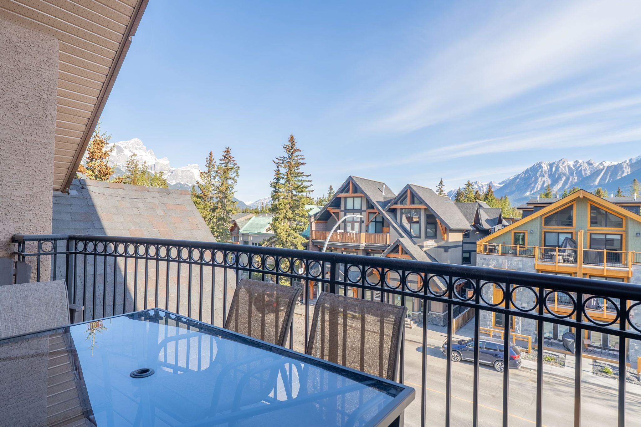 A balcony with a glass table and chairs, overlooking a street with cars and buildings, with mountains in the background.