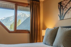 A hotel room with a bed, a window with curtains, a mountain view, and a wall-mounted lamp. - 3 Bedroom Penthouse  | Balcony + Fireplace (Canmore)