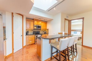 A modern kitchen with wooden cabinets, a black stove, and a glass island with bar stools. - 3 Bedroom Penthouse  | Balcony + Fireplace (Canmore)