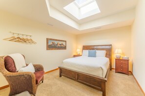 Een slaapkamer met dakraam, een bed met houten hoofdeinde, een rieten stoel, een nachtkastje met lampen, en een kapstok aan de muur.