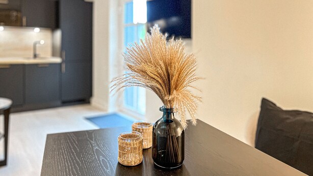 A modern kitchen with dark cabinetry, a dining table with a vase of dried wheat, and wicker candle holders. - Villa Triton Wohnung 08 (Ostseebad Boltenhagen)