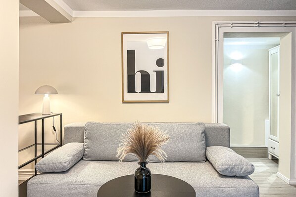 A modern living room with a grey sofa, a black coffee table, and a vase with dried plants. A framed 'hi' sign hangs on the wall, and a lamp is on a side table.