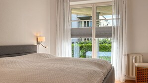 Une chambre à coucher avec un grand lit, une lampe de chevet, une fenêtre avec des rideaux, un radiateur et une chaise en bois.