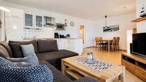 A modern living room with a sofa, coffee table, and a TV. There is a kitchen area with cabinets and a microwave. - Dünenblick Wohnung 39 (Ostseebad Boltenhagen)