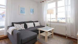 Un salón moderno con un sofá gris, una mesita pequeña y una lámpara de pie. Hay dos cuadros enmarcados en la pared y una ventana con cortinas blancas.