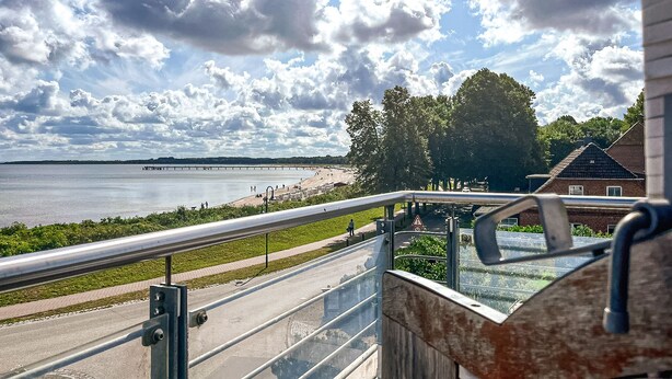 A view from a balcony overlooking a riverside promenade with a bridge in the distance, a pedestrian path, and a brick building. - Dünenblick Wohnung 17 (Ostseebad Boltenhagen)