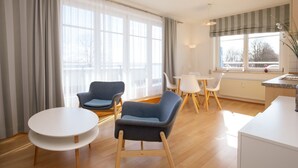 A modern living room with a round white table, two blue armchairs, a small white table, and a kitchen area with a striped window valance. - Dünenblick Wohnung 17 (Ostseebad Boltenhagen)
