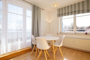 Ein Essbereich mit einem runden Tisch, vier Stühlen, großen Fenstern mit Vorhängen und einer Heizung.