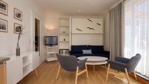 A modern living room with a sofa, two armchairs, a coffee table, and a TV on a stand. There are framed pictures on the wall and a window with curtains. - Dünenblick Wohnung 17 (Ostseebad Boltenhagen)