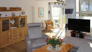 Un salon avec un canapé gris, une table basse en bois sur laquelle est posé un vase de fleurs, un téléviseur sur un meuble et un buffet en bois avec des portes en verre.