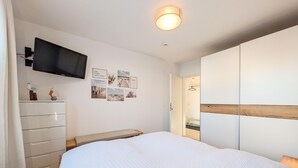 A bedroom with a bed, a TV mounted on the wall, a dresser, and a door leading to a bathroom. - Die Ferienwohnung Nr (Ostseebad Boltenhagen)