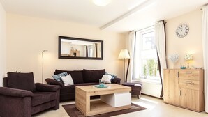 A modern living room with a dark brown sofa, a wooden coffee table, a large mirror, a wooden cabinet, and a window with curtains. - Die Ferienwohnung Nr (Ostseebad Boltenhagen)