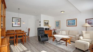 Ein geräumiges Wohnzimmer mit einem Holzesstisch, Stühlen, einem schwarzen Ledersessel, einem beigen Sofa und einem Couchtisch. An den Wänden hängen gerahmte Bilder, es gibt einen Holzschrank und eine Lampe auf einem Beistelltisch.