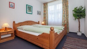 Ein hölzernes Bett mit zwei Kissen und einem Nachttisch mit Lampe. An der Wand hängen zwei gerahmte Bilder und neben dem Fenster steht ein Blumentopf.