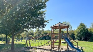 Un parque infantil con un tobogán de madera y columpios, rodeado de césped y árboles.
