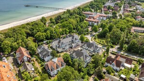 Vista aerea di una località costiera con spiaggia, molo e edifici residenziali immersi nel verde.