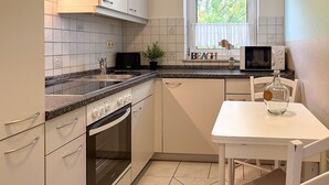 Et lille køkken med hvide skabe, indbygget ovn, mikroovn og et vindue med udsigt til grønne omgivelser.