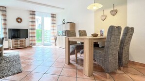 A spacious living area with a wooden dining table, cushioned chairs, a television on a stand, and a large window with curtains. - Sünnslag Wohnung 105 (Ostseebad Boltenhagen)