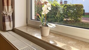 A window with a potted plant and a view of a house and garden. - Sünnslag Wohnung 105 (Ostseebad Boltenhagen)