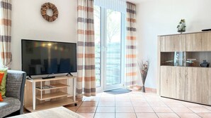 A living room with a flat-screen TV on a wooden stand, a sofa, a coffee table with decorative items, a cabinet with glass doors, and a view through a sliding glass door. - Sünnslag Wohnung 105 (Ostseebad Boltenhagen)