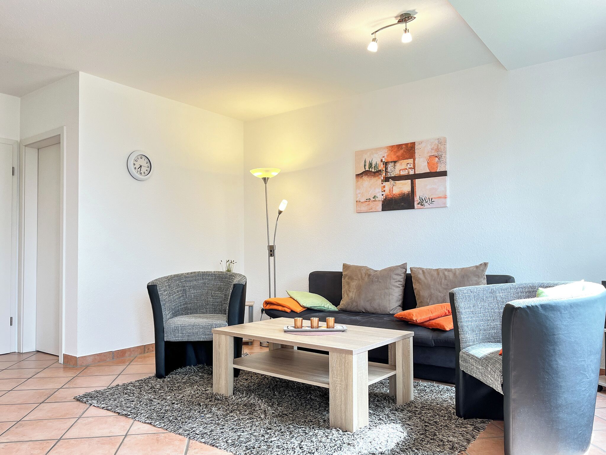 Ein modernes Wohnzimmer mit einer Couch, zwei Sesseln, einem Couchtisch, einer Stehlampe und einer Wanduhr.