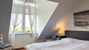 Un dormitorio con cama, ventana con cortinas translúcidas, radiador, silla, mesita de noche con lámpara y un cuadro en la pared.