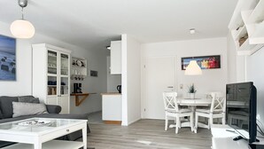 Een moderne woonkamer met een glazen salontafel, een witte bank, een eethoek met een witte tafel en stoelen, en een keuken met witte kasten en een wit aanrecht.