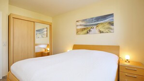 A neatly made bed with a white duvet, a wooden headboard, and bedside lamps. There is a wooden wardrobe with a mirror and a painting of a beach scene on the wall.