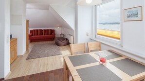 Ein modernes Wohnzimmer mit einem roten Sofa, einem Esstisch mit Unterlagen, einem Sitzsack und Blick auf das Meer durch ein großes Fenster.