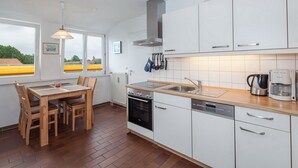 Eine moderne Küche mit weißen Schränken, einer Edelstahl-Dunstabzugshaube, einem Geschirrspüler und einer Kaffeemaschine. Es gibt einen Esstisch mit Stühlen und ein Fenster mit Blick auf ein gelbes Gebäude.