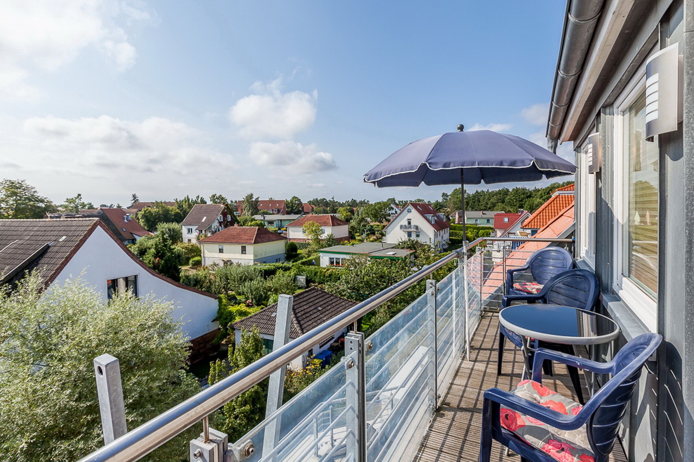 Een balkon met een tafel en stoelen, een grote parasol en uitzicht op huizen en groen.