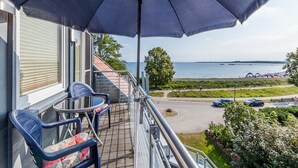 Ein Balkon mit blauem Sonnenschirm, zwei Stühlen und einem Tisch mit Blick auf ein Gewässer und eine Straße mit Autos.