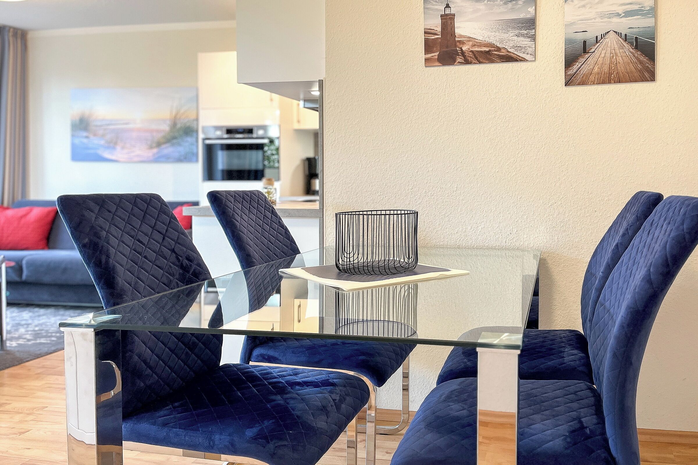 Ett modernt vardagsrum med ett matbord med glasskiva, omgivet av blå sammetsstolar, ett kök i bakgrunden och en målning av en fyr på väggen.